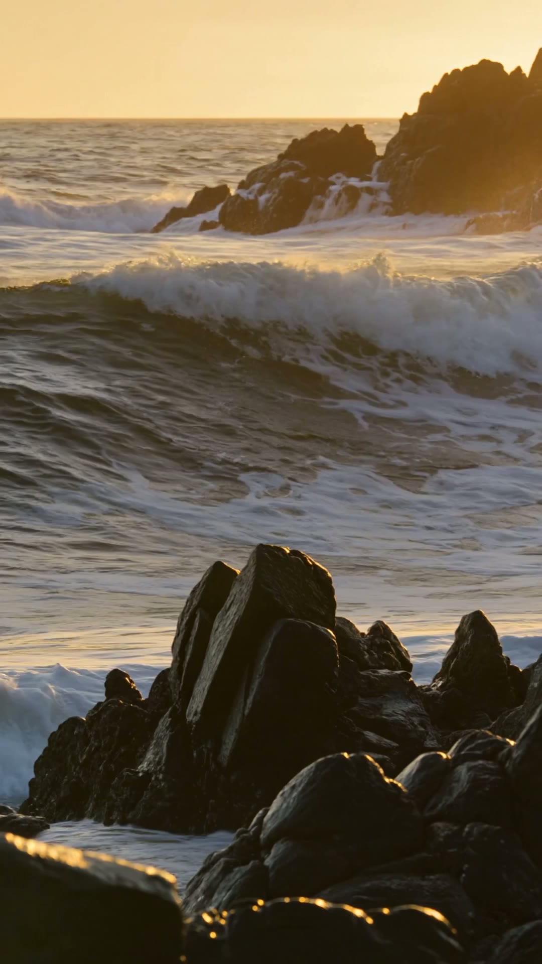 Powerful ocean waves crashing against dark jagged rocks at sunset, dramatic spray and…